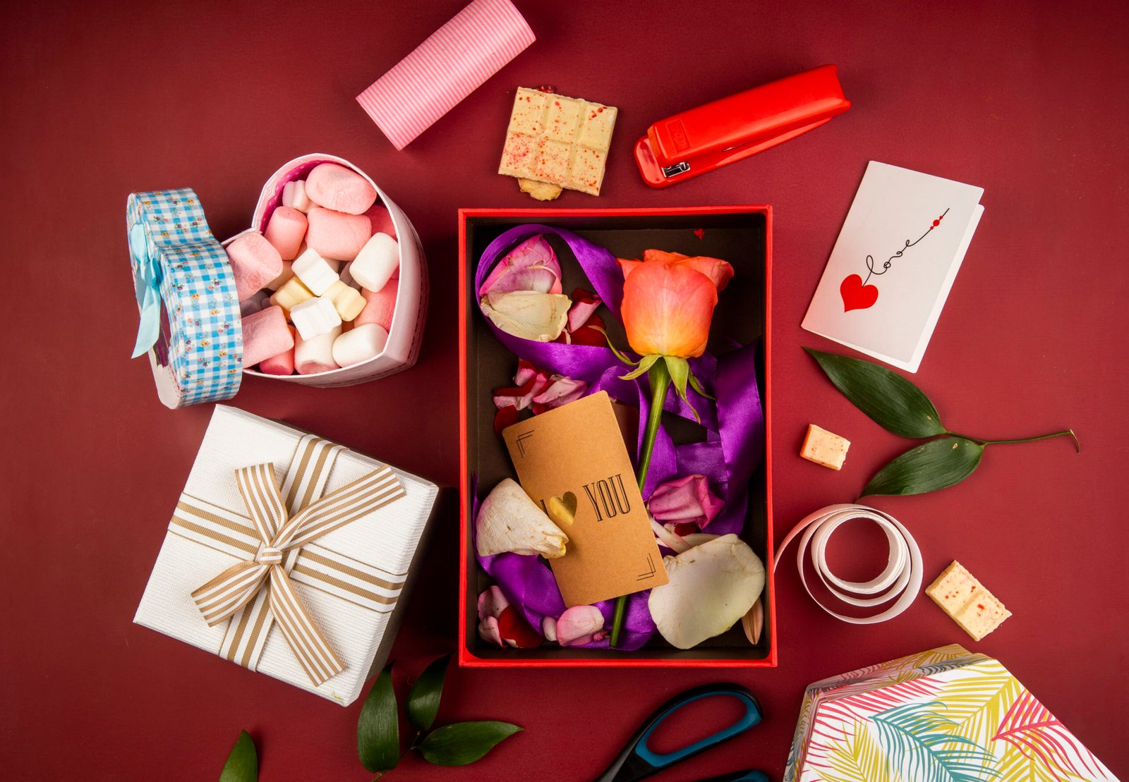 top view of a red present box with brown paper card and coral color rose flower and petals with purple ribbon and heart shaped box filled with marshmallow on dark red background